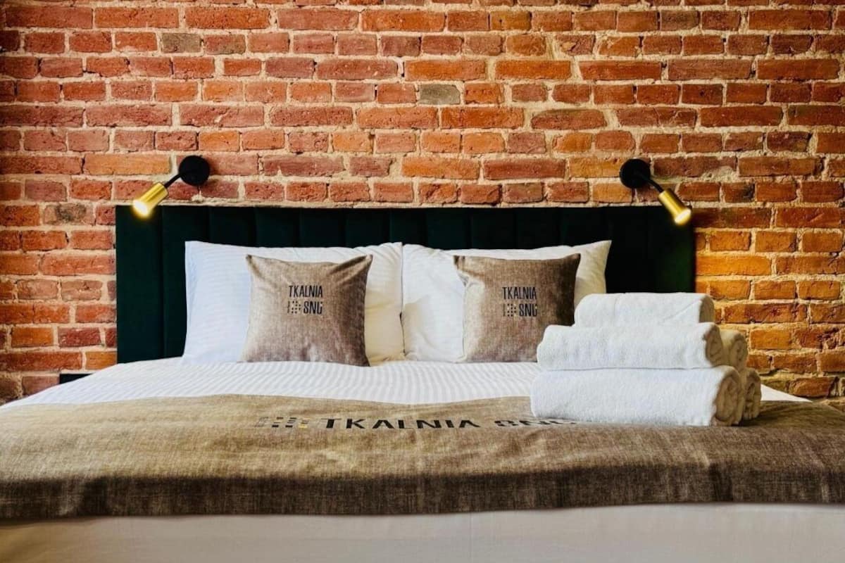 Una cama bien hecha con un cabecero verde, ropa blanca de cama y almohadas con el texto "TIKALNIA". Hay dos lámparas de pared a cada lado de la cama.