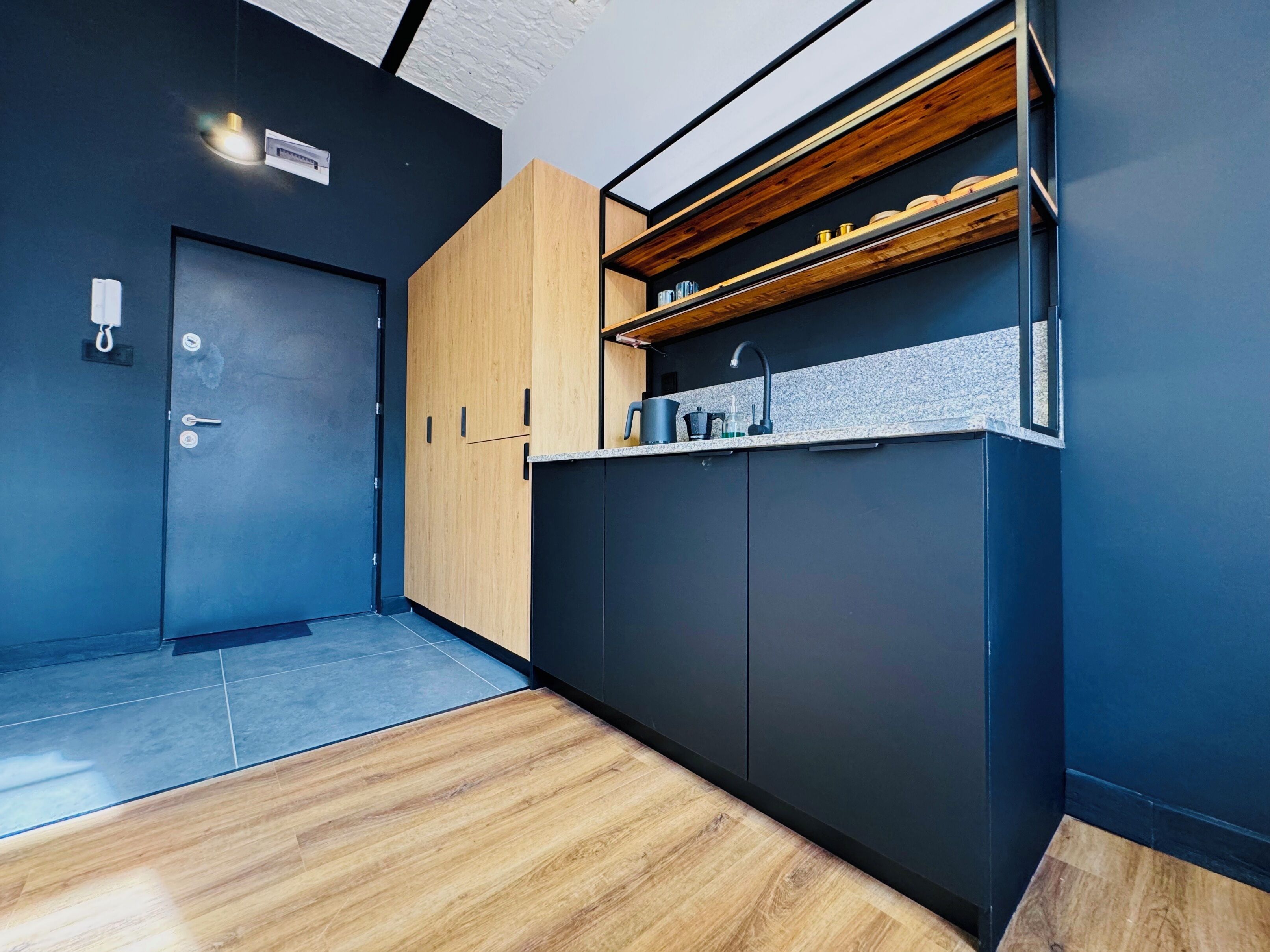 A modern kitchen with dark cabinetry, a wooden door, and a countertop with a sink and kettle.