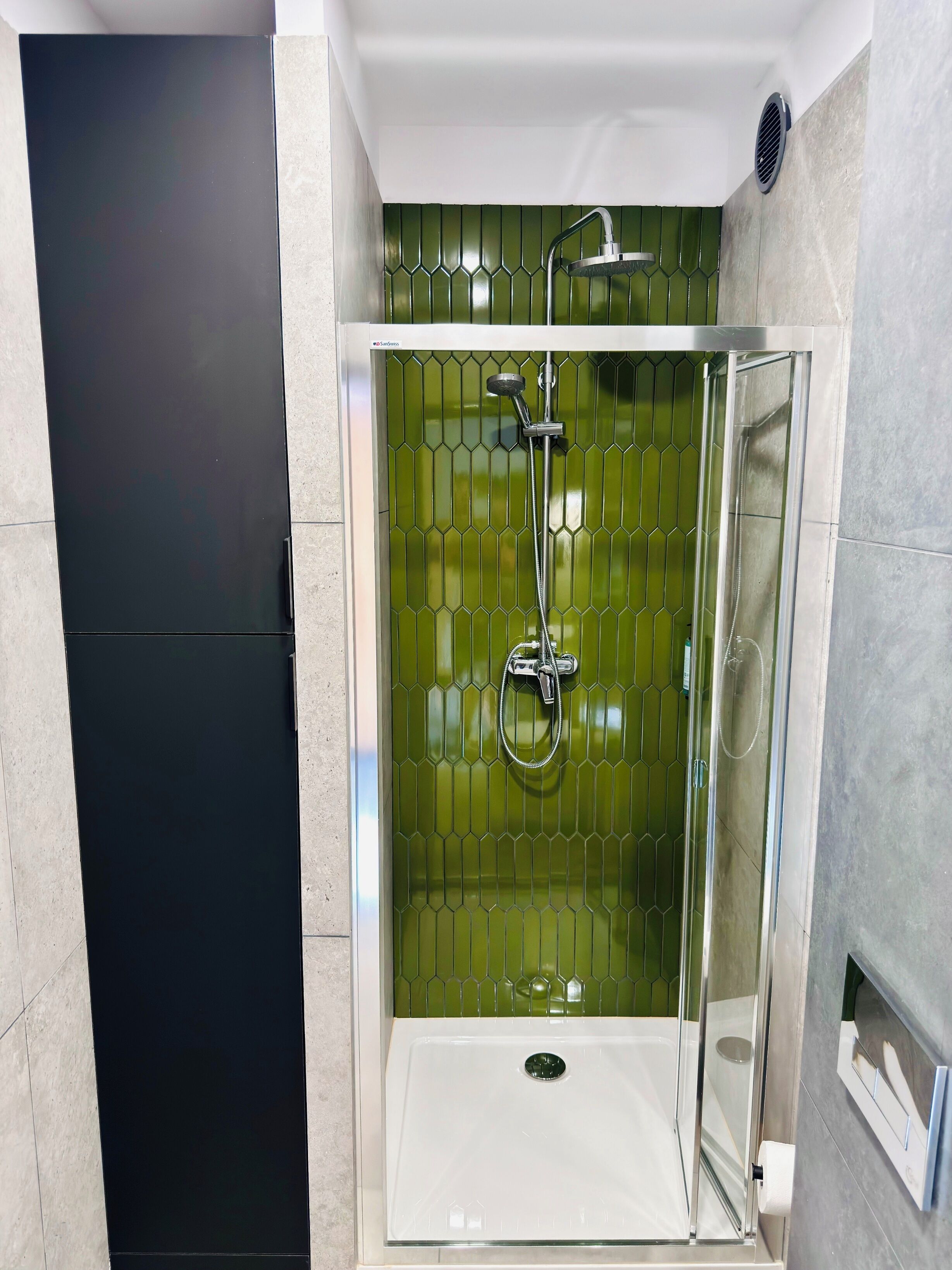 Une douche fermée par des parois de verre, avec un mur recouvert de carreaux verts, un lavabo blanc et des toilettes.