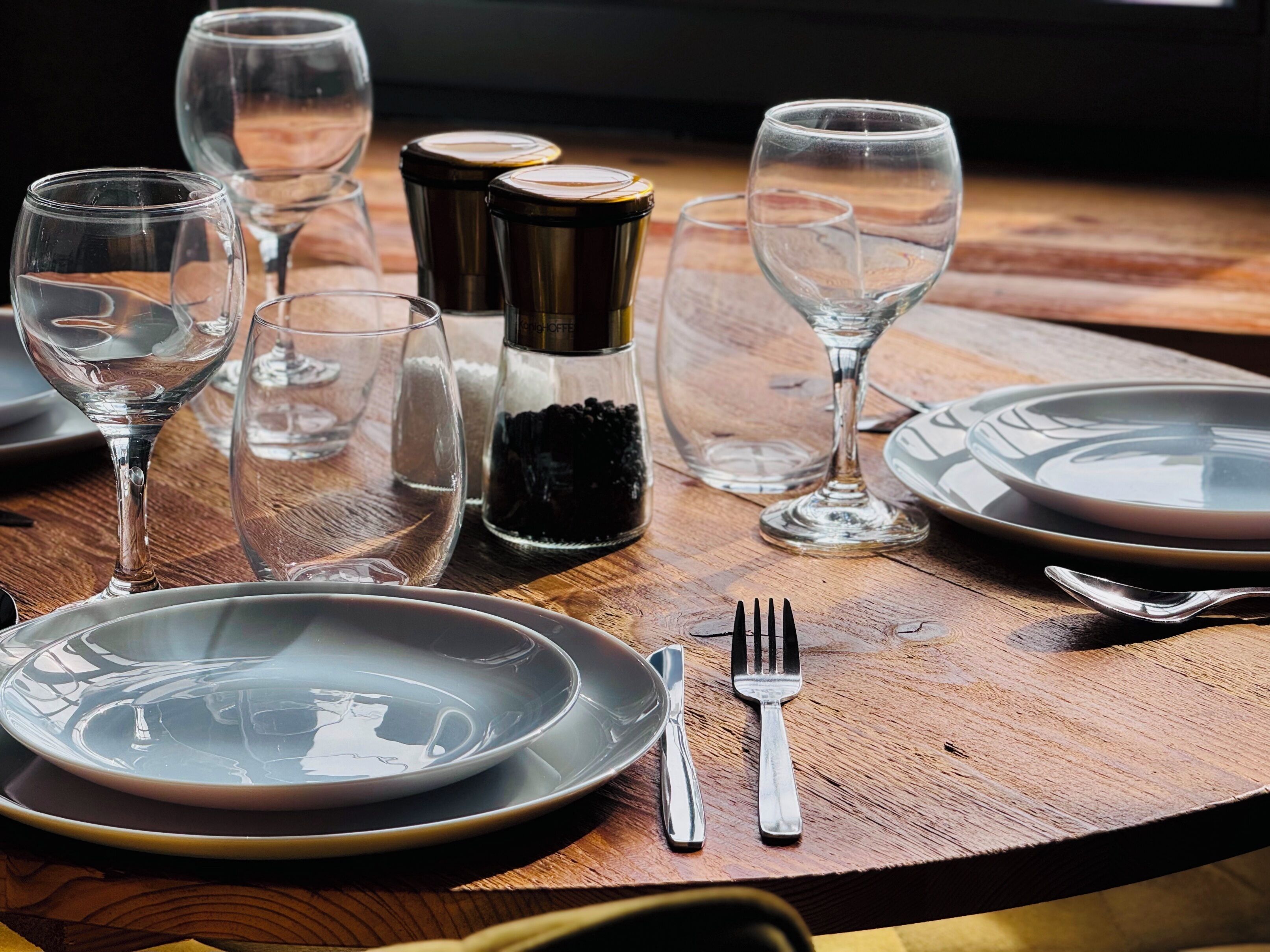 Una mesa de madera dispuesta con platos, cubiertos y copas de vino vacías.