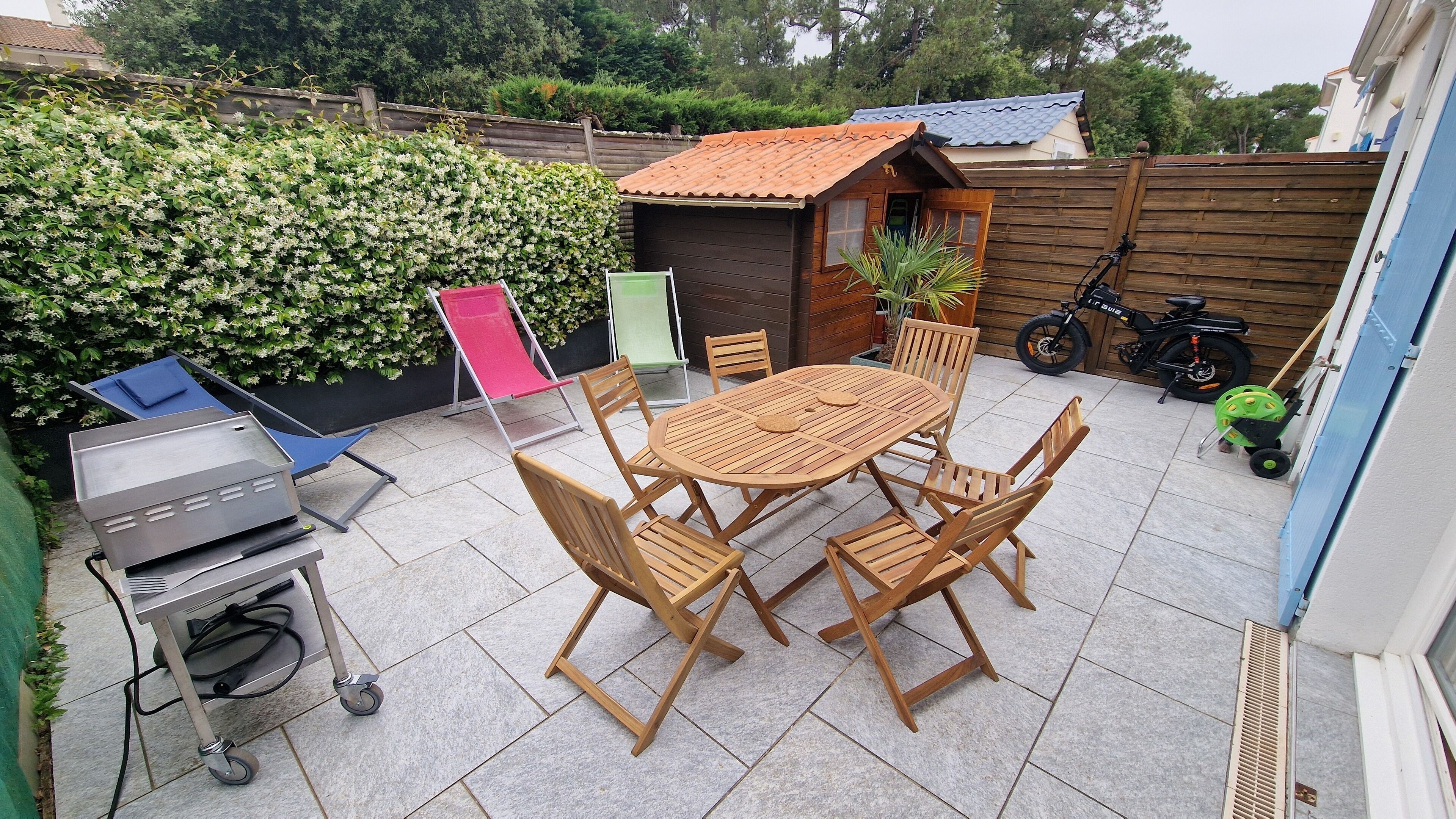 Un patio con una mesa y sillas de madera redondas, una parrilla, una silla plegable y un cobertizo pequeño.