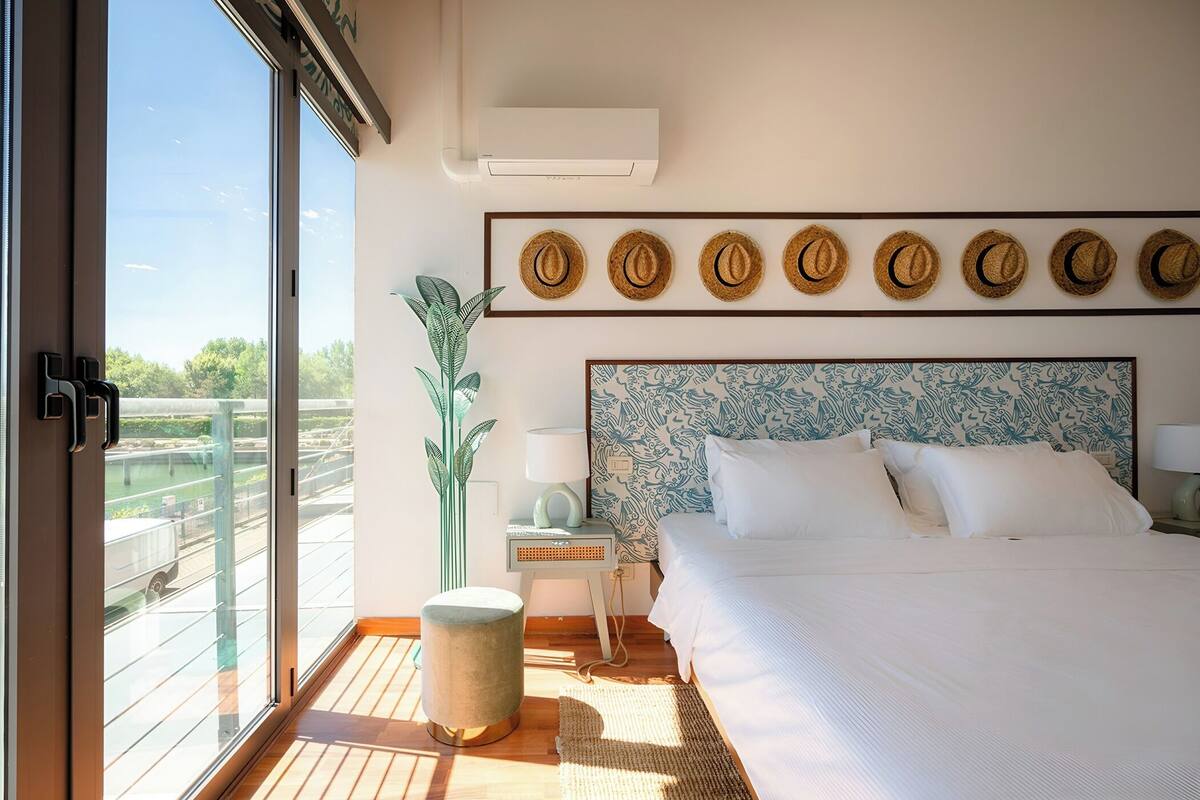 A hotel room with a large bed, a nightstand with a lamp, a wall-mounted air conditioner, a window with a view of greenery, and a series of hats on the wall.