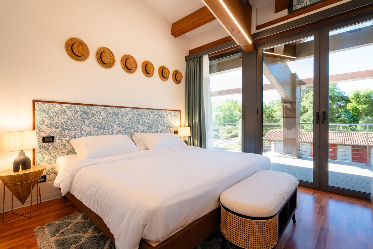 A bedroom with a large bed, a bedside table, a lamp, a wicker bench, and a view through a glass door.