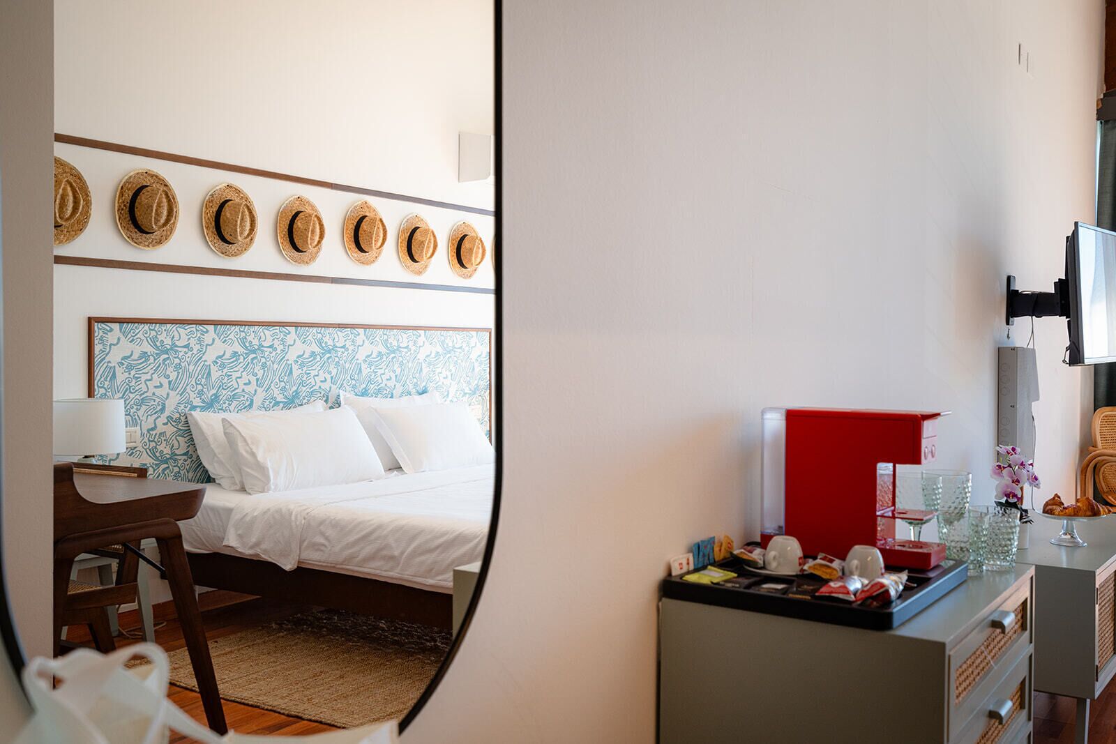 A hotel room with a bed, a desk, a chair, a small table with a red box, and a TV on the wall.