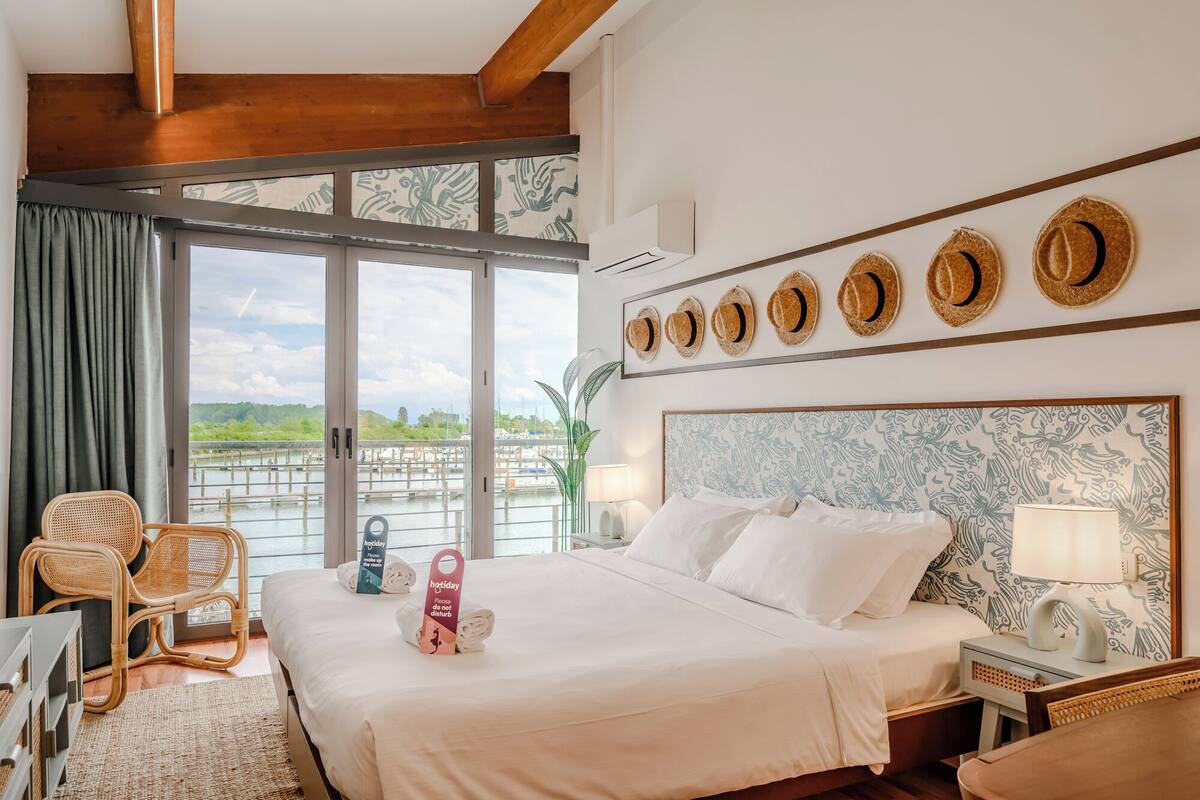 A hotel room with a bed, a desk, a chair, a view of a marina, and a large window.