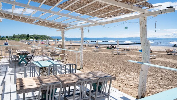 A beachside restaurant with outdoor seating, a wooden deck, and a view of the beach and boats in the background. - Hotiday Grado Marina (Grado)