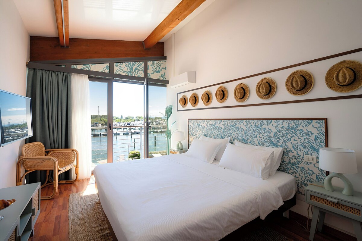 Ein Hotelzimmer mit einem großen Bett, einem Fernseher, einem Schreibtisch, Korbstühlen, einem Balkon mit Blick und einer Wand, die mit Hüten dekoriert ist.