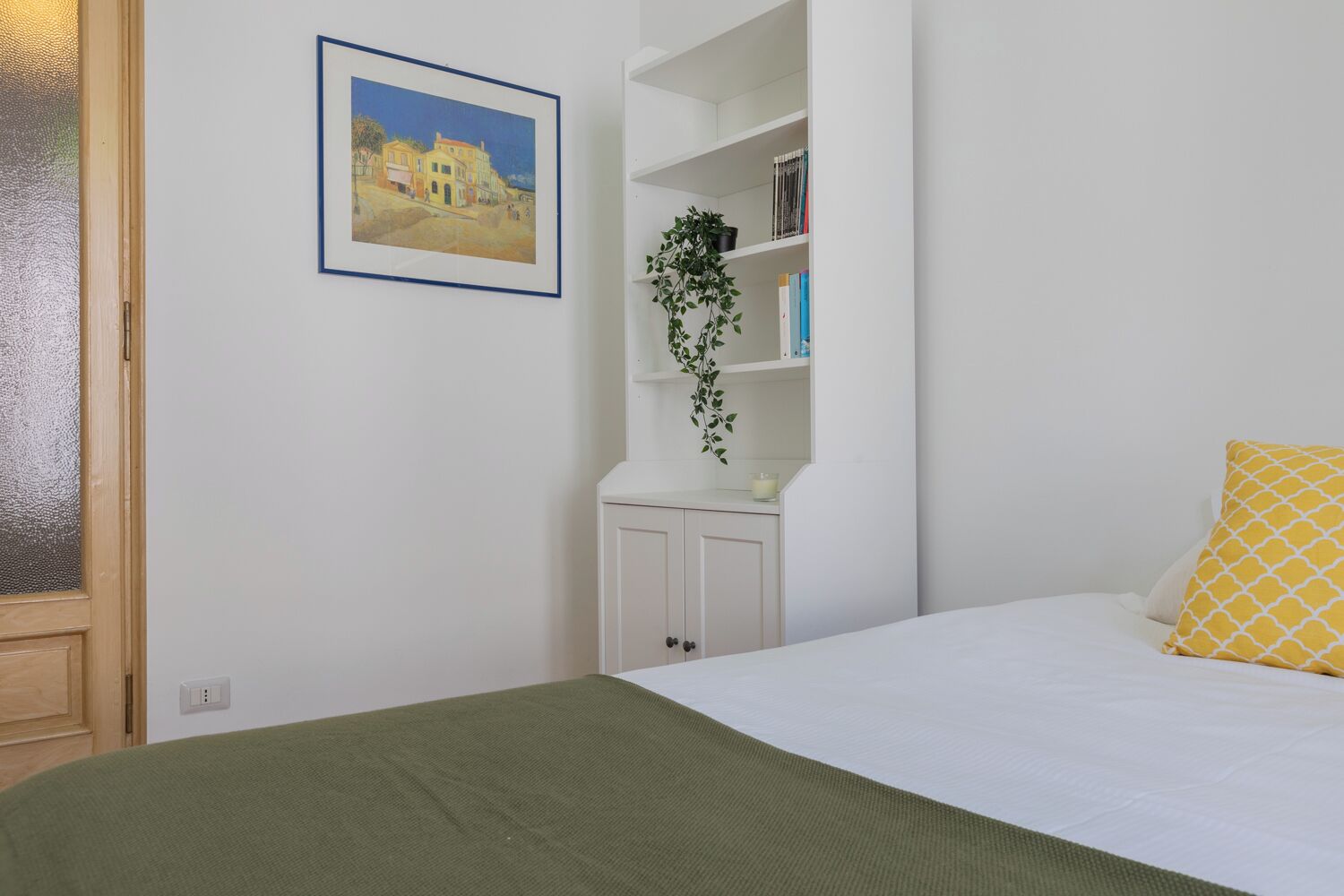 A bedroom with a bed, a green blanket, a yellow pillow, a white nightstand, a white shelf with books and a plant, and a framed painting on the wall.