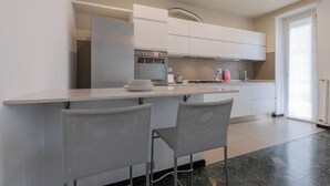 A modern kitchen with white cabinets, a stainless steel refrigerator, and a gas stove. There are two grey chairs at a small island counter. - Casa Gabri by Wonderful Italy (Turin)