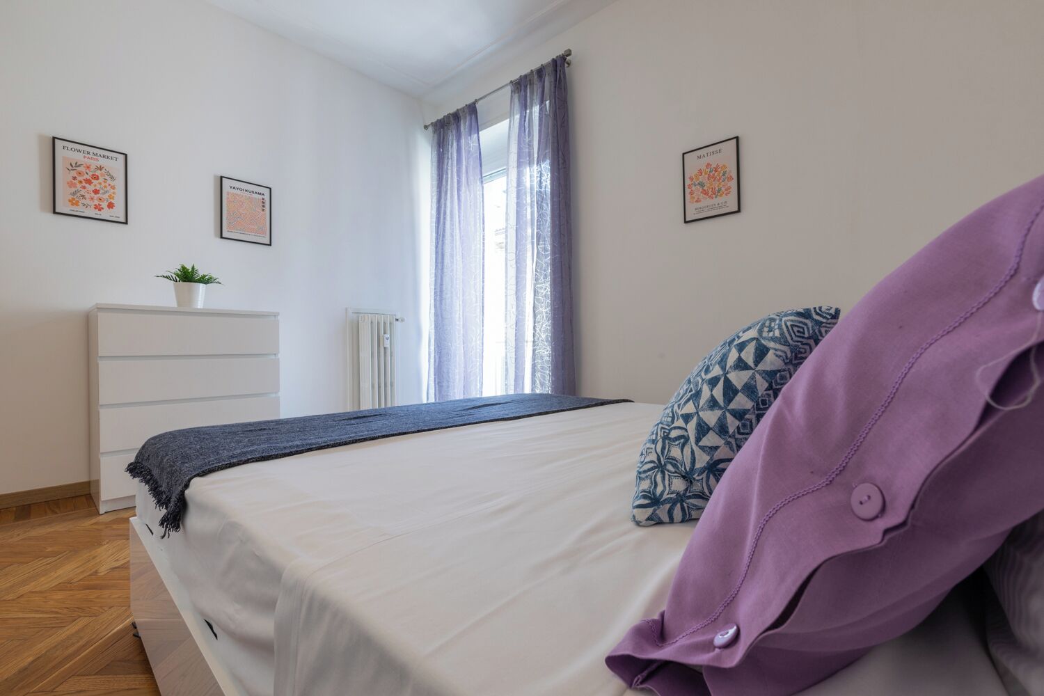 A bedroom with a bed, a dresser, a radiator, and two framed artworks on the wall.