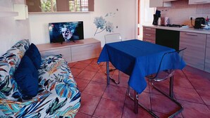 A living room with a sofa, a TV, a dining table with a blue tablecloth, and a kitchen area.