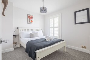 A bedroom with a white bed, grey bedding, a black and white pendant light, a framed picture on the wall, and a window with blinds. - Charming Central Cottage (Cambridge)