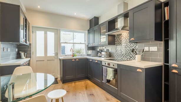 A modern kitchen with dark cabinets, a glass dining table, and a view of the outdoors. - Beautiful 2bed Central Home (Cambridge)