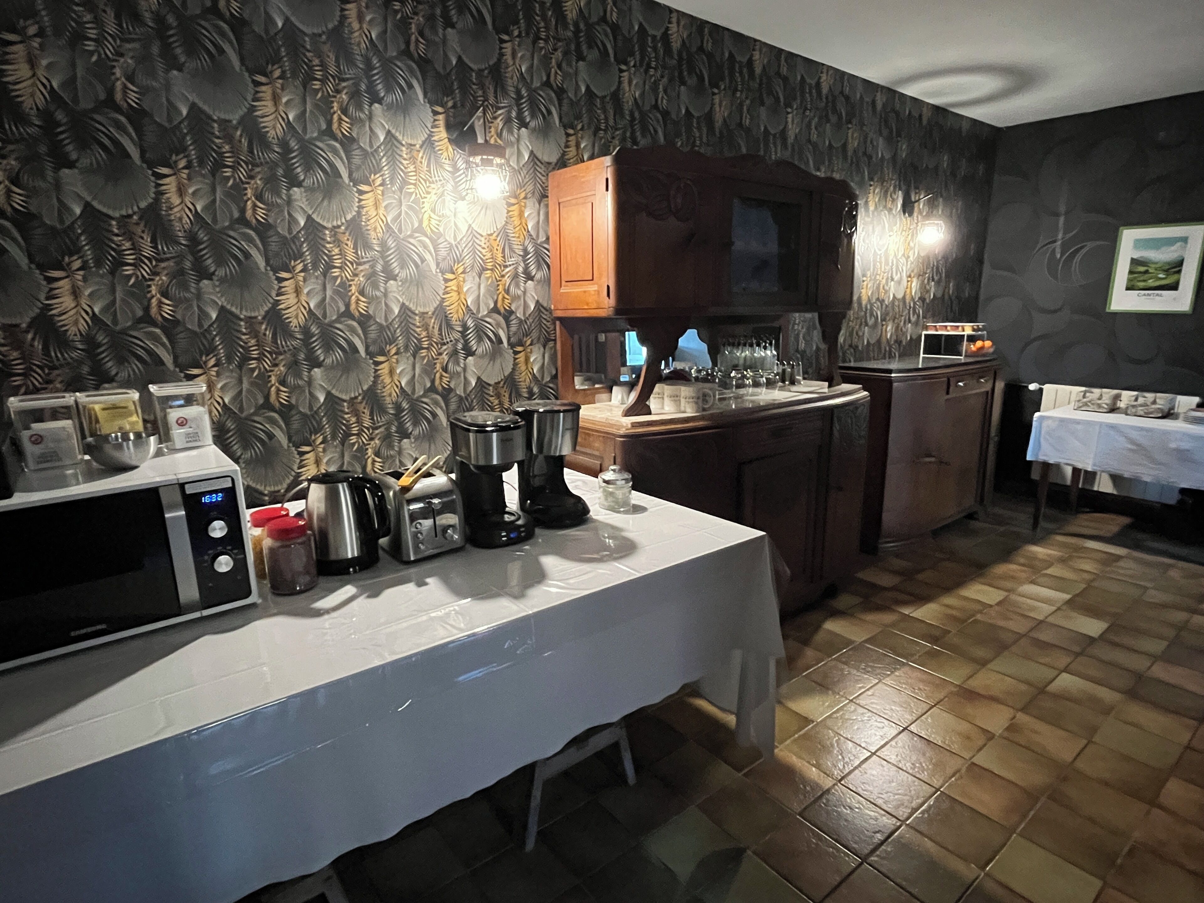 A kitchen with a microwave, toaster, coffee maker, and kettle on the counter. There is a wooden cabinet above the counter and a table with a white tablecloth.