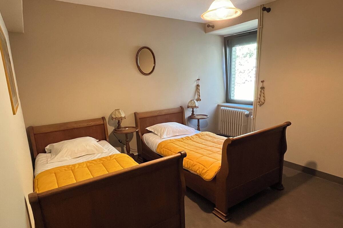 Una habitación con dos camas de madera, cada una con colcha amarilla, una mesita de noche con lámpara, un espejo redondo en la pared y una ventana con cortinas.