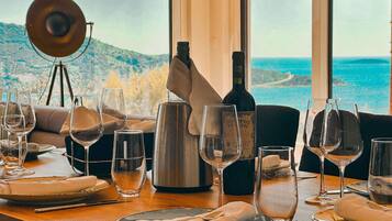 A dining table set with wine glasses, a bottle of wine, and a metal pitcher, with a scenic view of the sea and hills in the background.