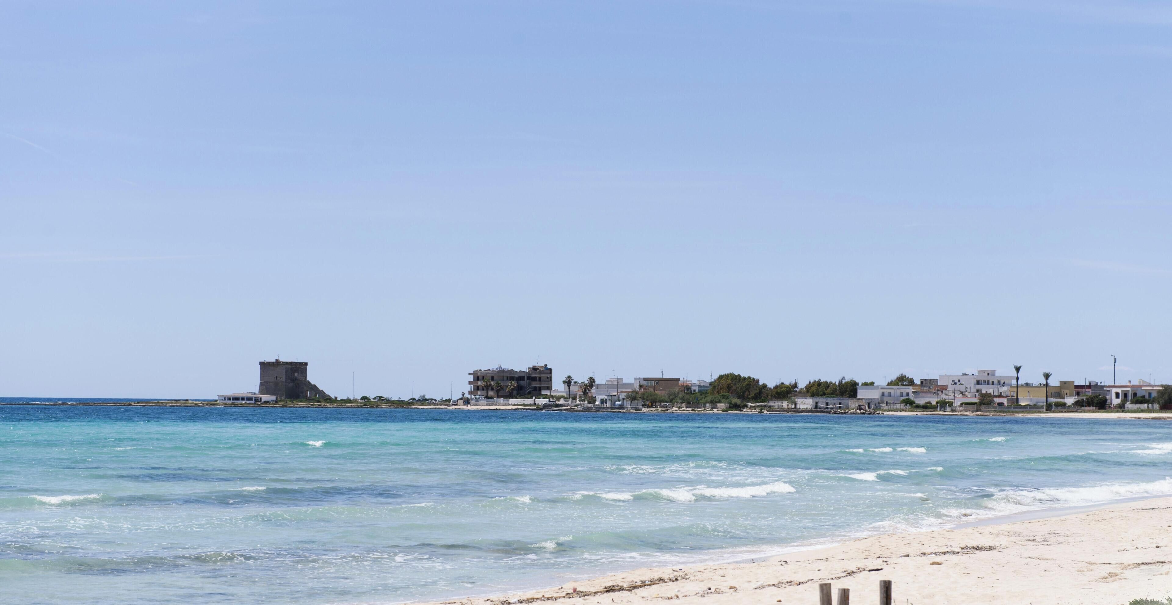 Una playa con agua azul cristalina, una orilla arenosa y una hilera de edificios al fondo.