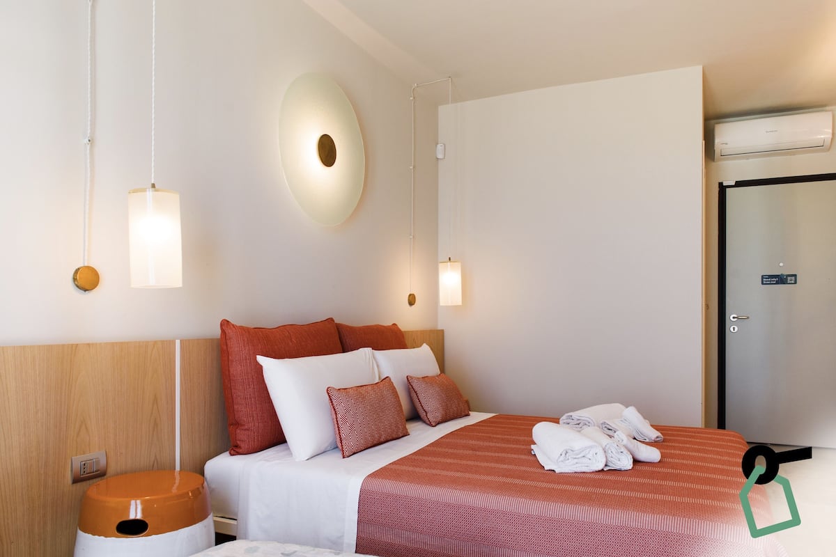 Una habitación de hotel con cama, almohadas, mesita de noche, lámpara de pared y puerta.