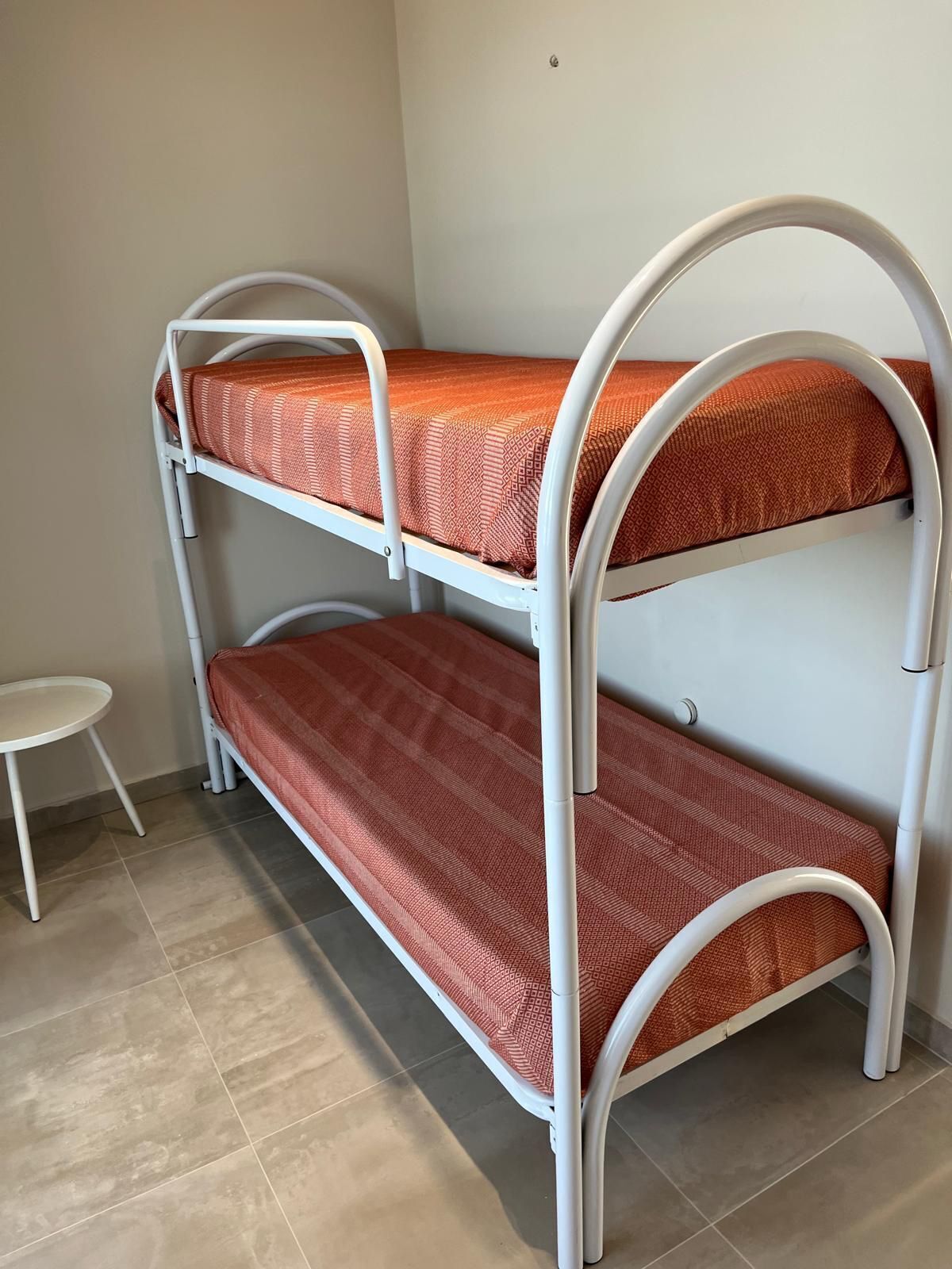 Una cama litera de dos niveles, una mesita blanca pequeña y una pared lisa.