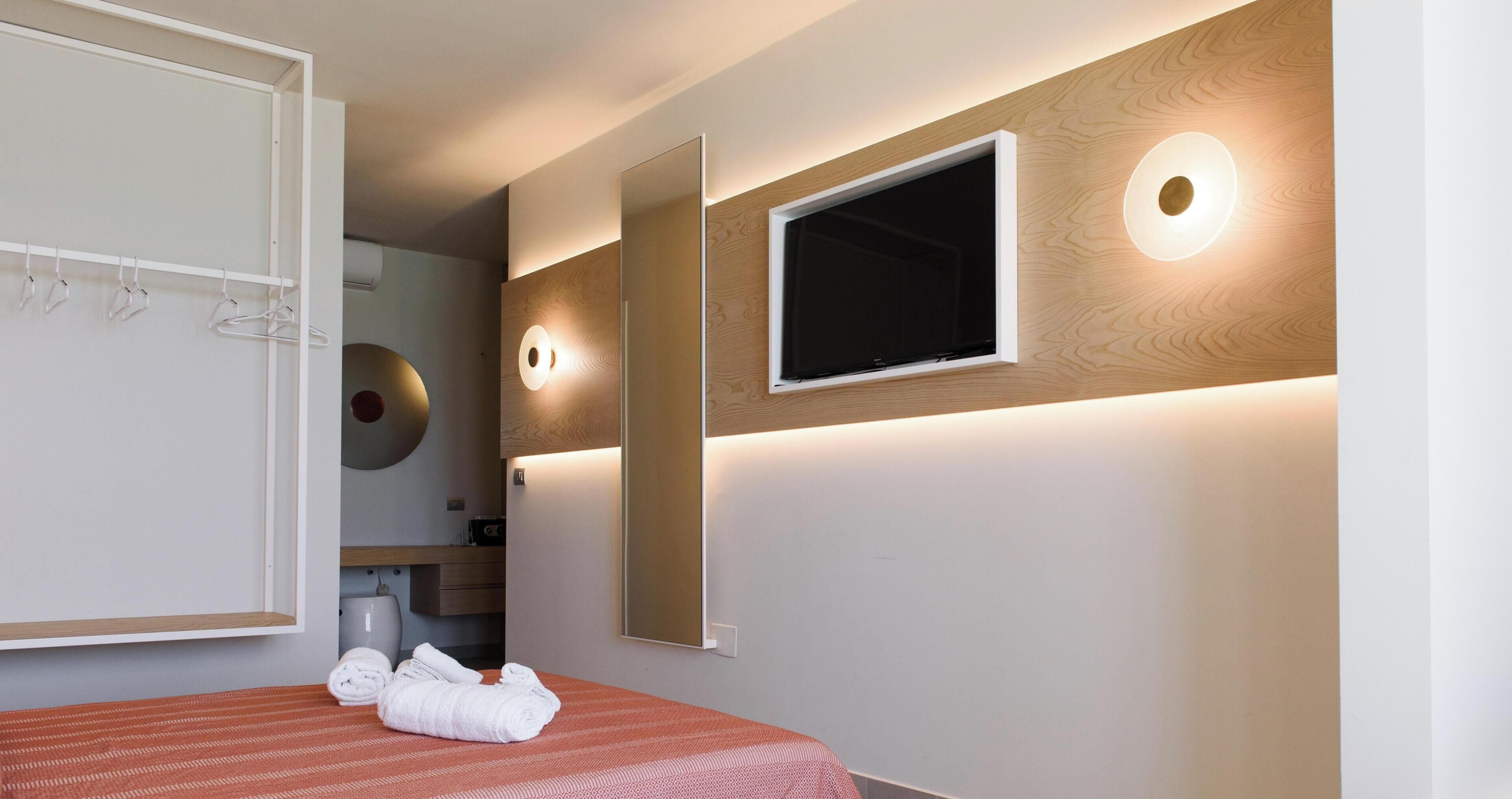 A hotel room with a bed, a mirror, a television, and a wall-mounted light fixture.