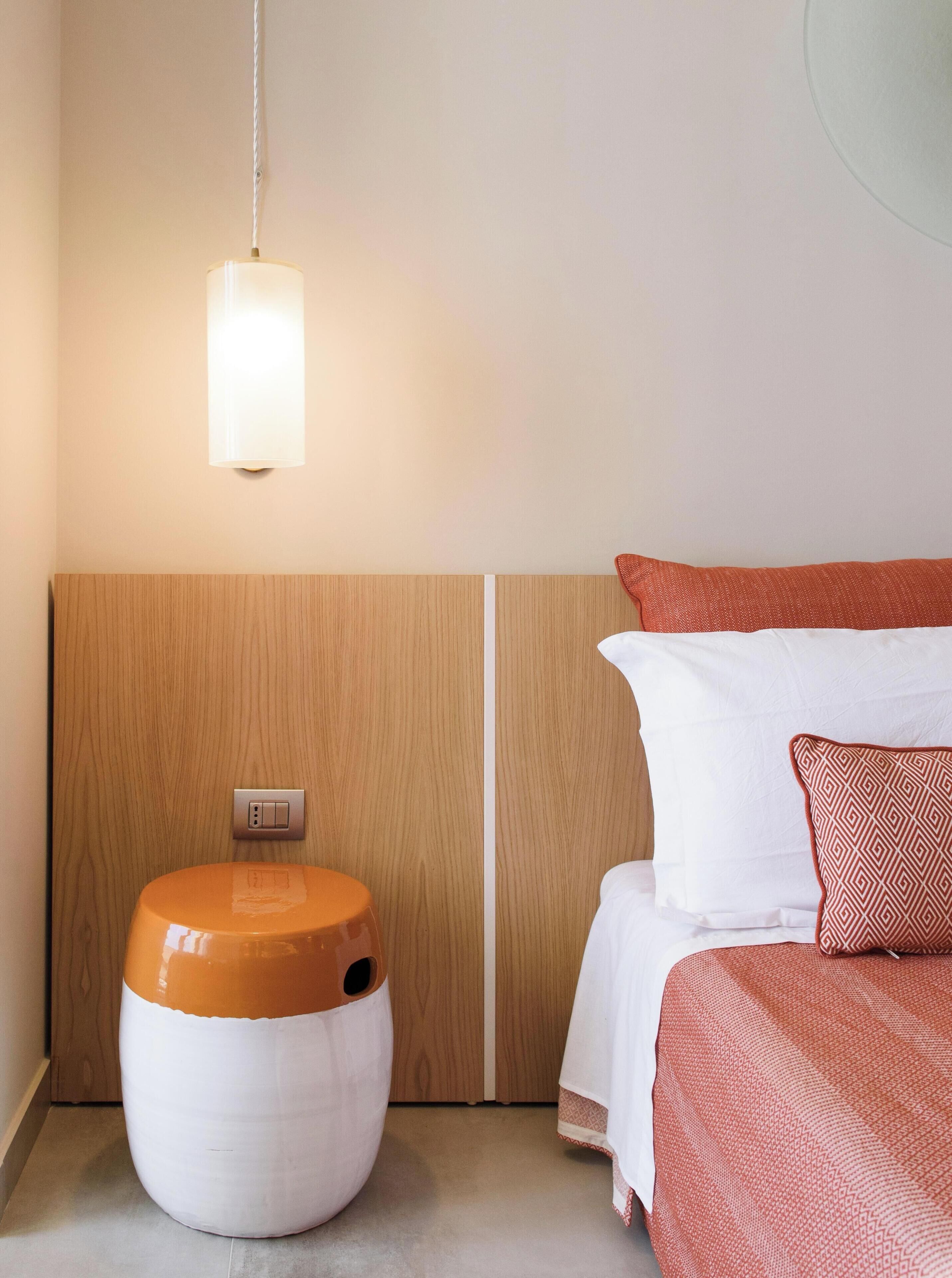 Habitación de hotel con cabecera de madera, mesita de noche con lámpara, cama con ropa de cama estampada y toma de corriente en la pared.