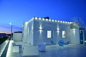 Un edificio moderno con techo plano, zona de estar exterior con sillas blancas y azules, y una escalera que lleva a un nivel superior.