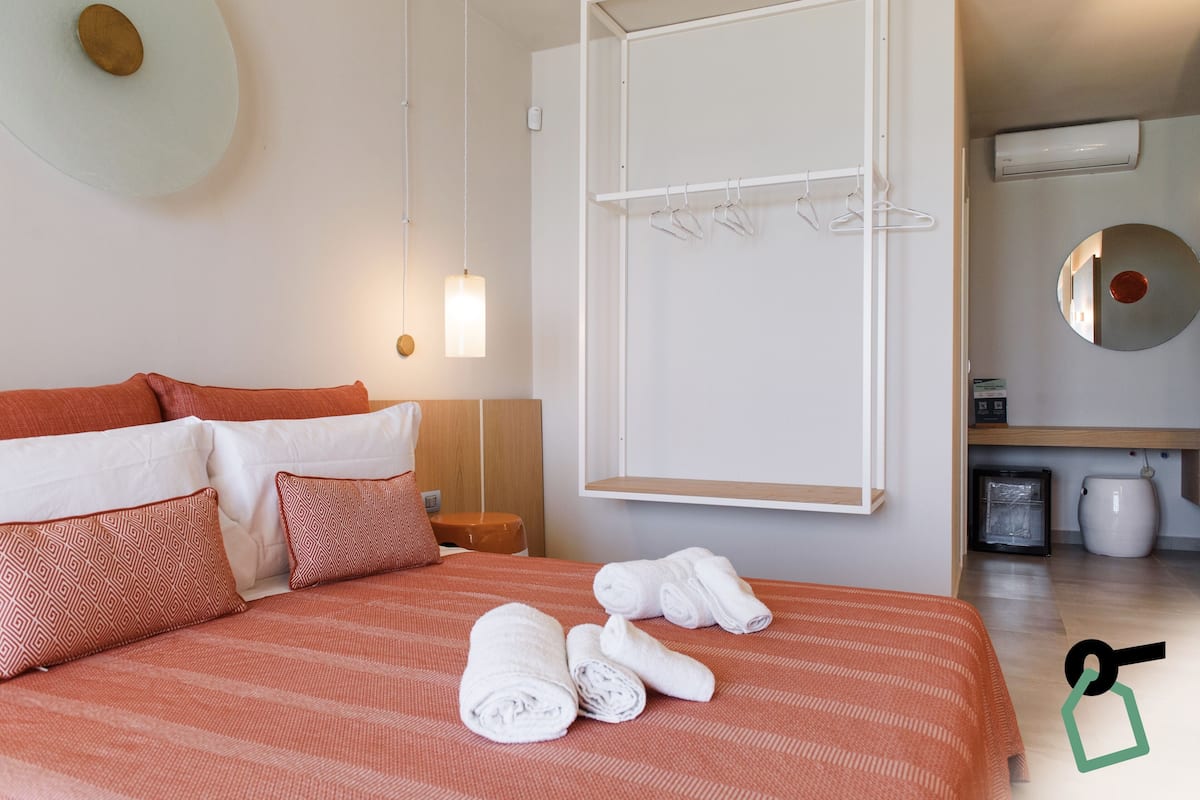 Una cama bien hecha con una colcha naranja a rayas, almohadas blancas y toallas enrolladas de color blanco. Hay una mesita de noche de madera con una lámpara, una pared blanca con un espejo circular y una pizarra blanca.