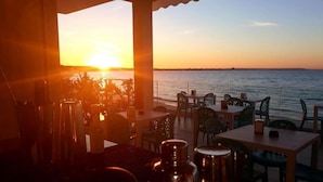 Een buitenzitplaats met tafels en stoelen gericht op een zonsondergang boven het water.