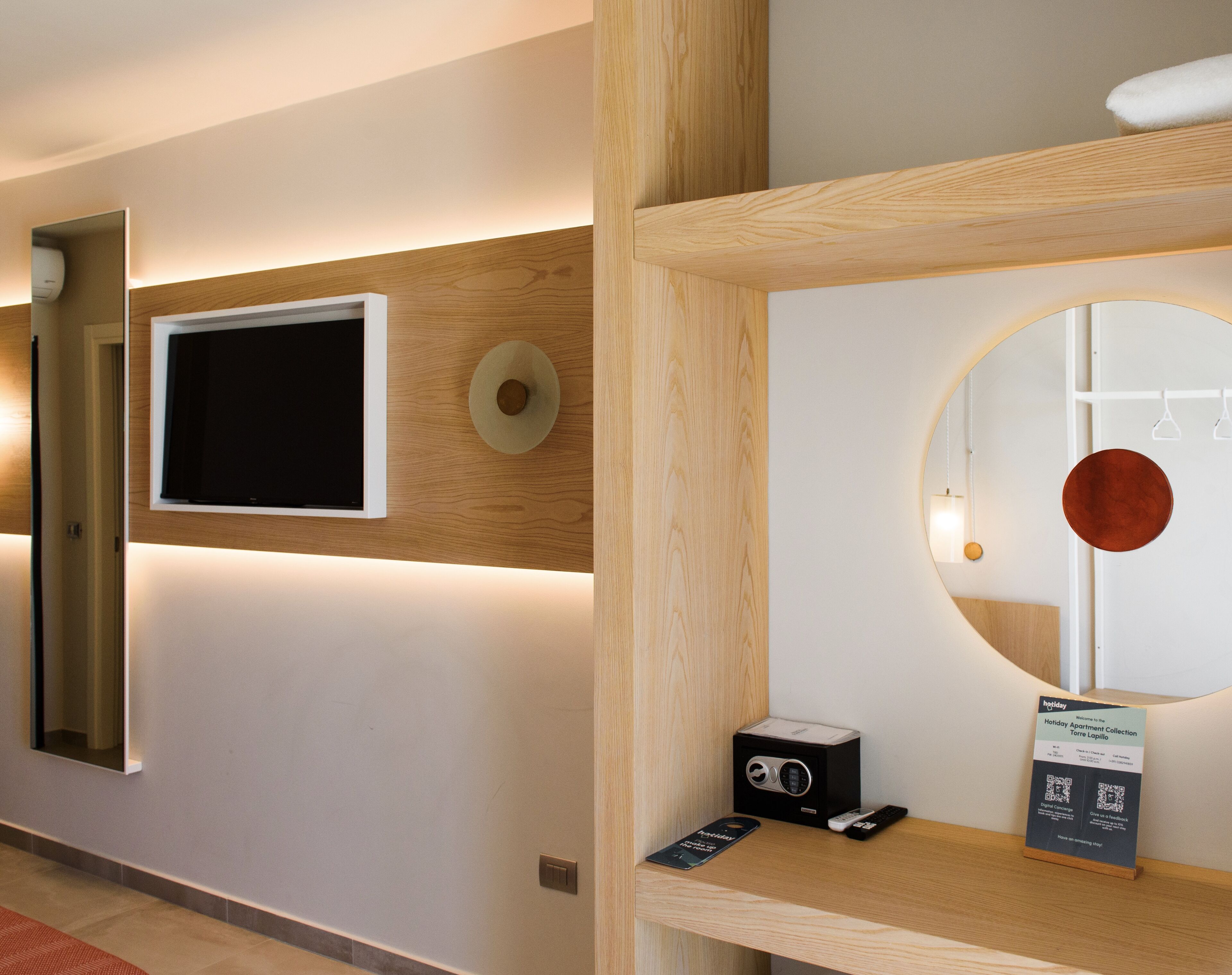 Una hall d'albergo moderna con una TV a schermo piatto, uno specchio rotondo, una reception in legno e un cartello per Holiday Apartment Collection.