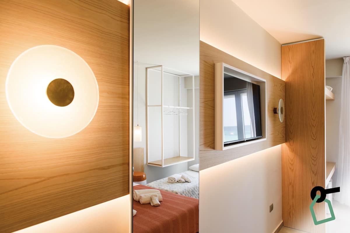 Una habitación de hotel moderna con suelo de madera, una cama con manta roja, una mesita de noche blanca y un espejo grande.
