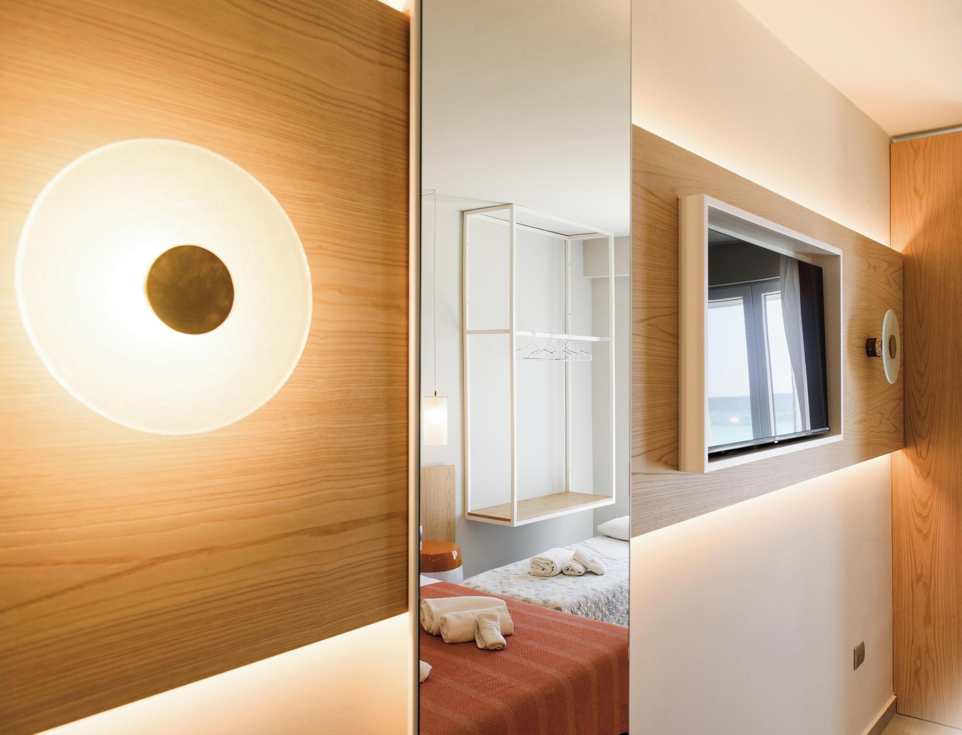 Ein modernes Hotelzimmer mit einer großen, runden Leuchte, einem Kleiderschrank, einem Bett mit roter Decke und einem Fenster mit Vorhängen.