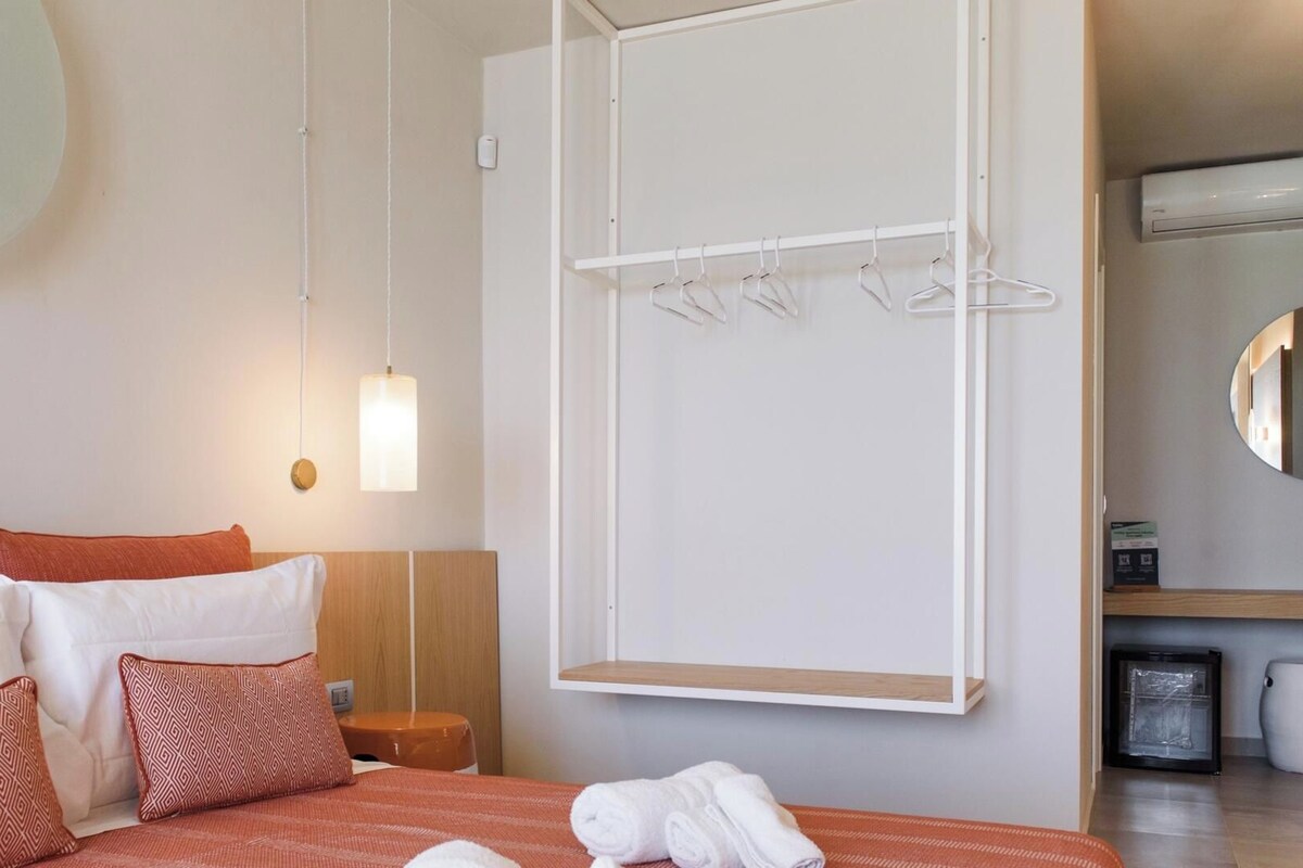 Een hotelkamer met een bed, nachtkastje, hanglamp, een handdoekhouder aan de muur en een spiegel.