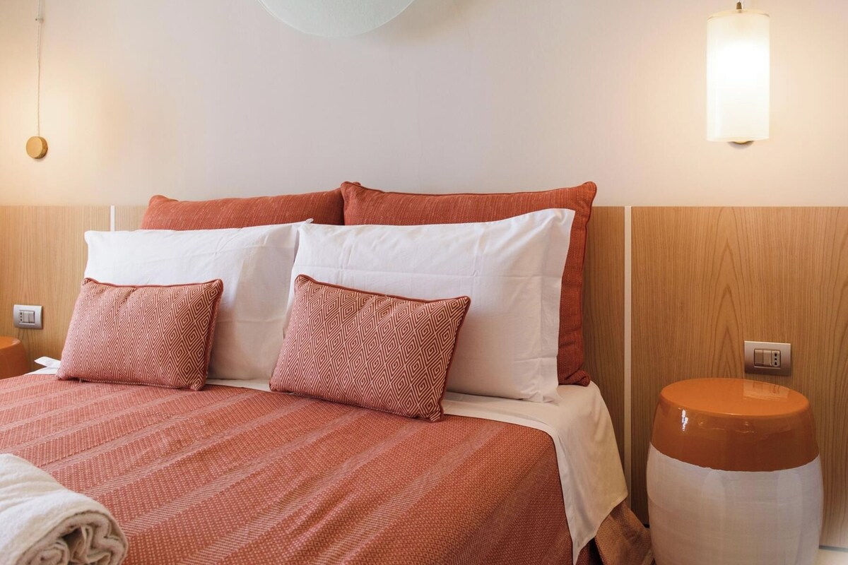 Habitación de hotel con cama, almohadas, mesita de noche, lámpara y aplique de pared.