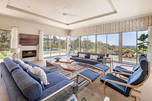 A spacious living room with a large sofa, lounge chairs, and a coffee table. There is a television mounted on the wall and a large window offering a view of the outdoors. - PICTURESQUE RURAL OASIS ON 6 ACRES BOASTS PANORAMIC BUSHLAND AND OCEAN VIEWS (Quindalup)