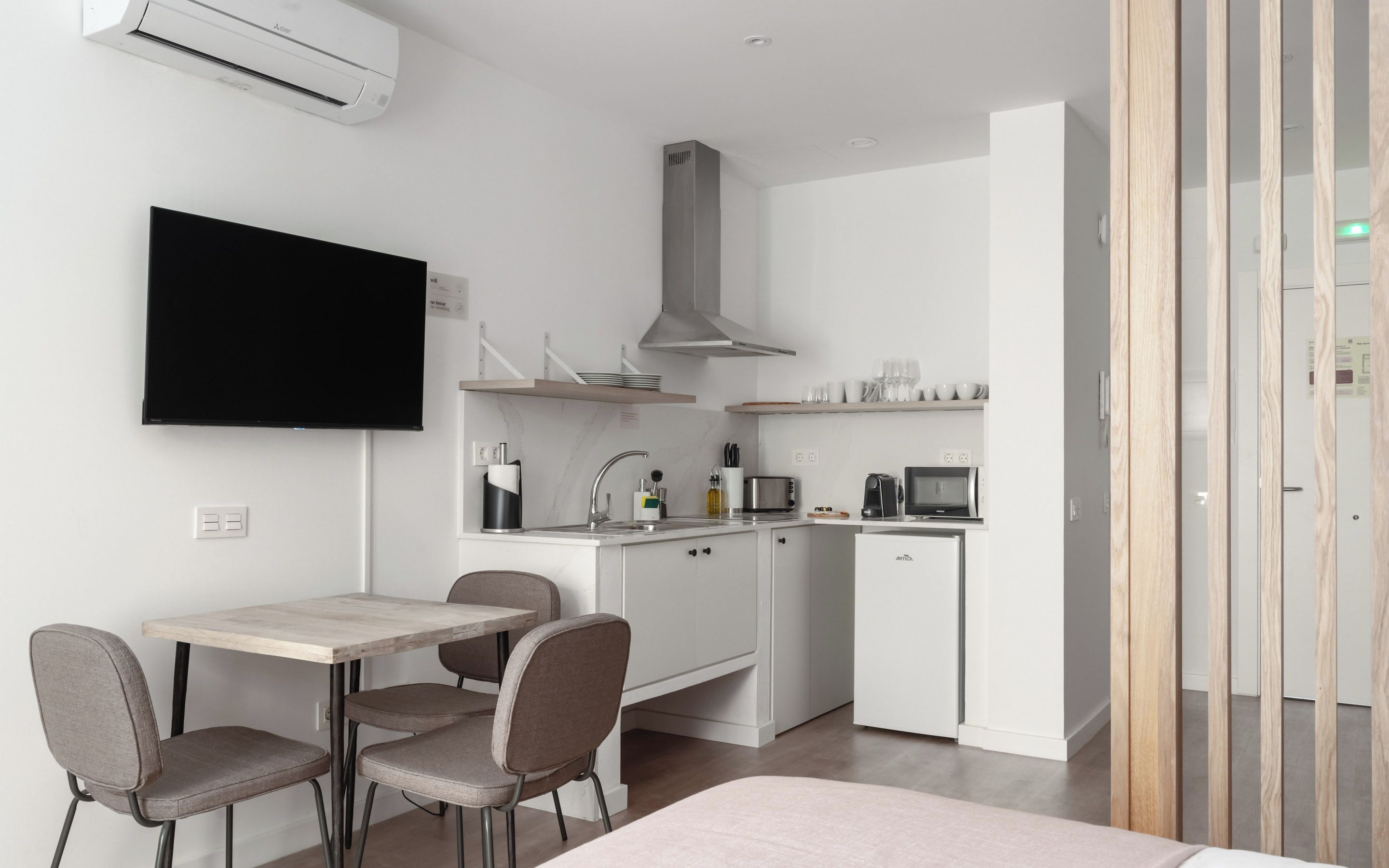 A modern kitchen with white cabinets, a stainless steel range hood, and a microwave. There is a dining area with a table and chairs, and a flat-screen TV mounted on the wall.