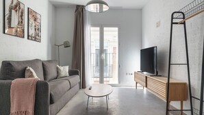 A modern living room with a grey sofa, a small round coffee table, a TV on a wooden stand, and a balcony with a metal railing. - Limehome Almeria C. Hernán Cortés (Almeria)