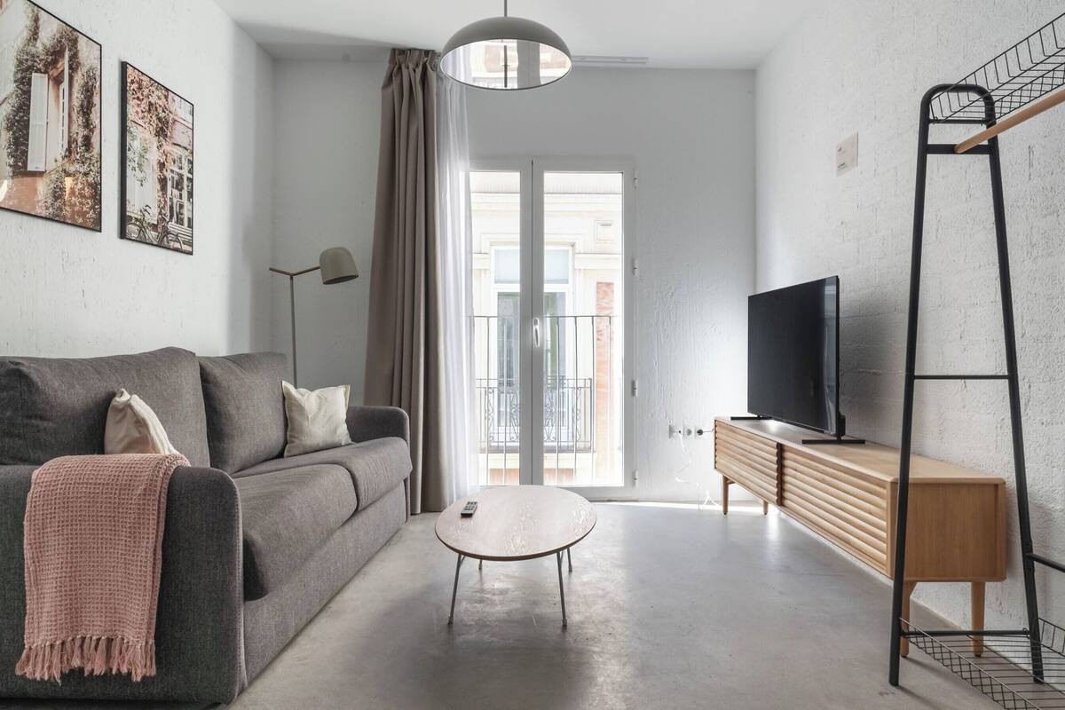 Ein modernes Wohnzimmer mit einer grauen Couch, einem kleinen runden Couchtisch, einem Fernseher auf einem Holzgestell und einem Balkon mit Metallgeländer.