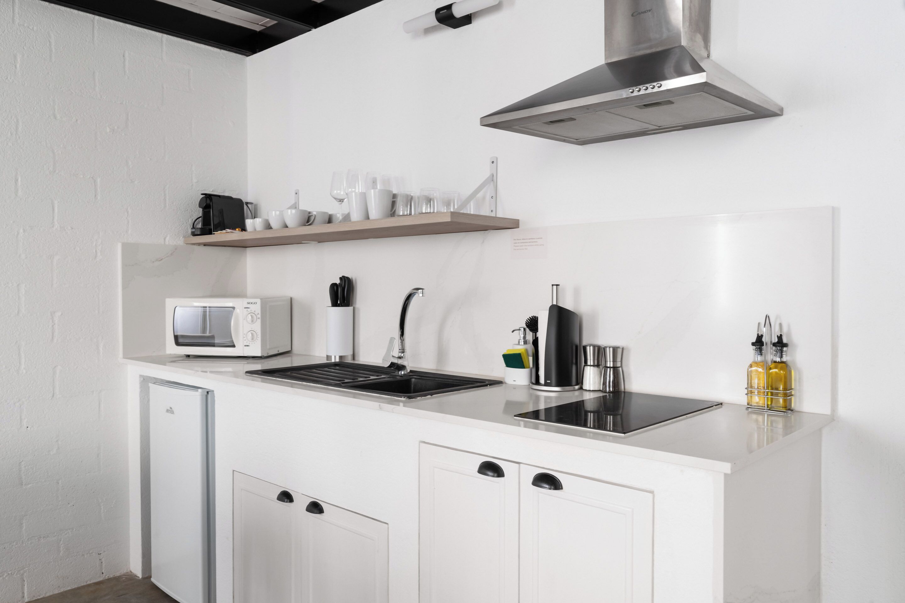 Una cocina compacta con microondas integrado, un fregadero y una campana extractora de acero inoxidable. Hay un refrigerador pequeño y un estante con tazas y vasos.
