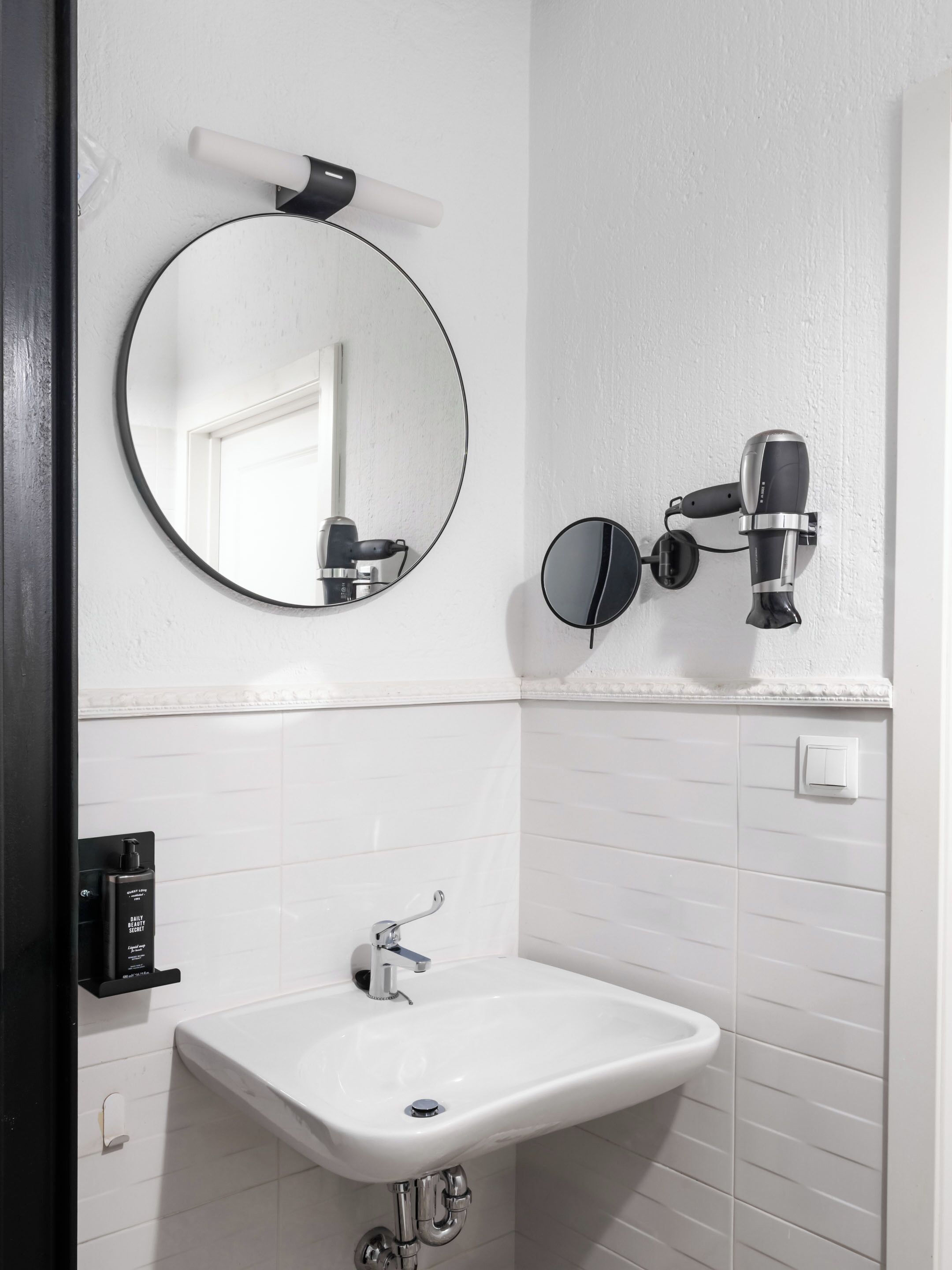 Un baño con un espejo redondo, un secador de pelo montado en la pared y un lavabo blanco con grifo cromado.