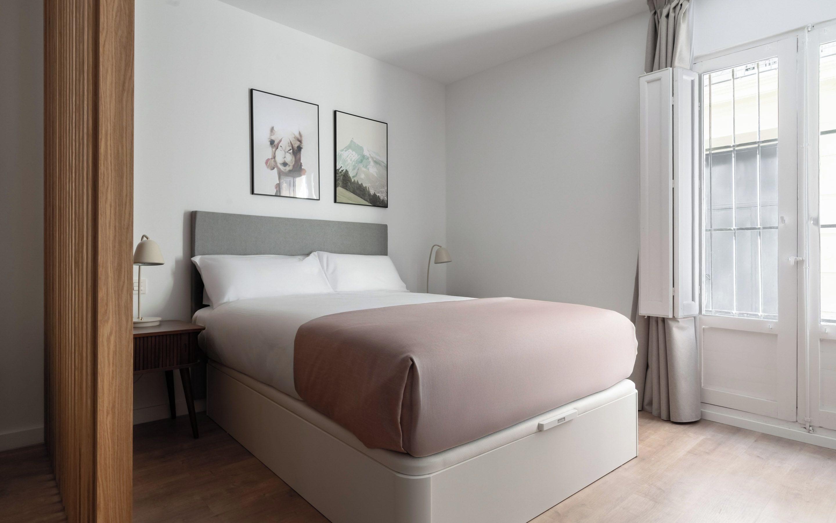 Een moderne slaapkamer met een bed, nachtkastje, lamp en twee foto’s in lijsten aan de muur.