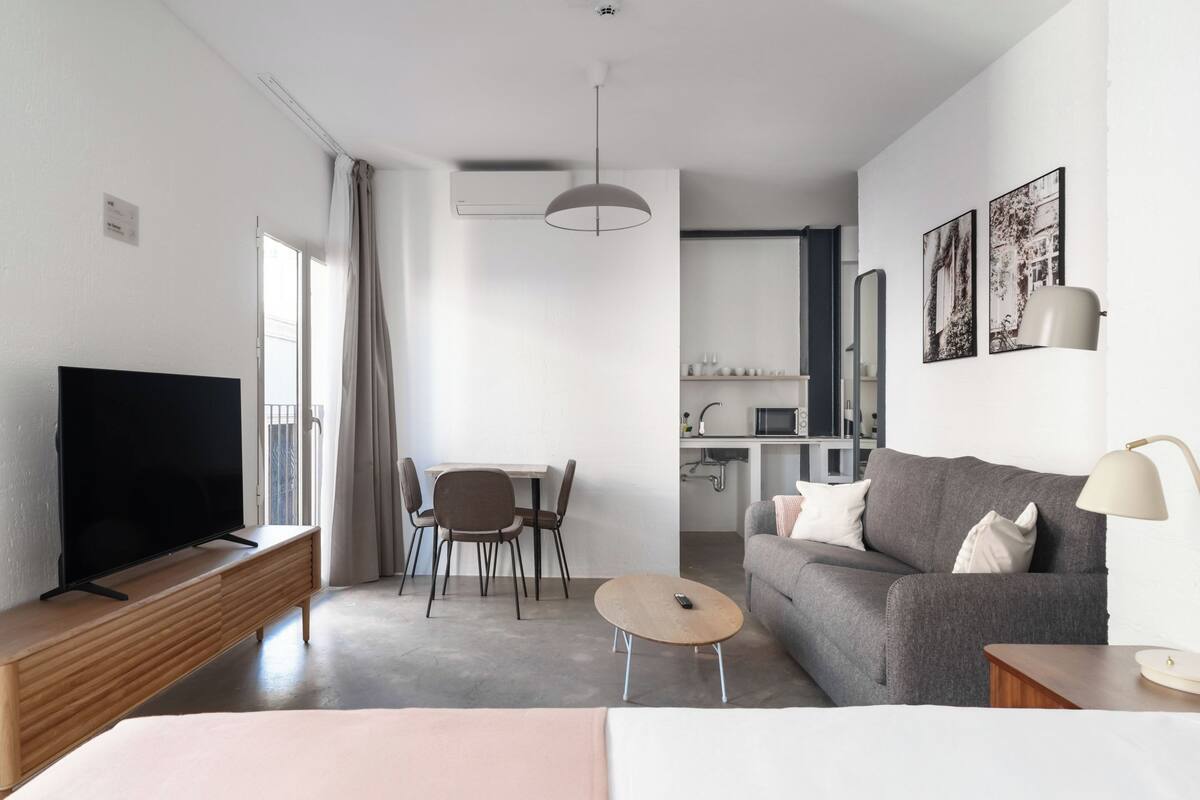 Ein modernes Wohnzimmer mit Flachbildfernseher, einem grauen Sofa, einem kleinen runden Couchtisch und einem Essbereich mit Tisch und Stühlen.