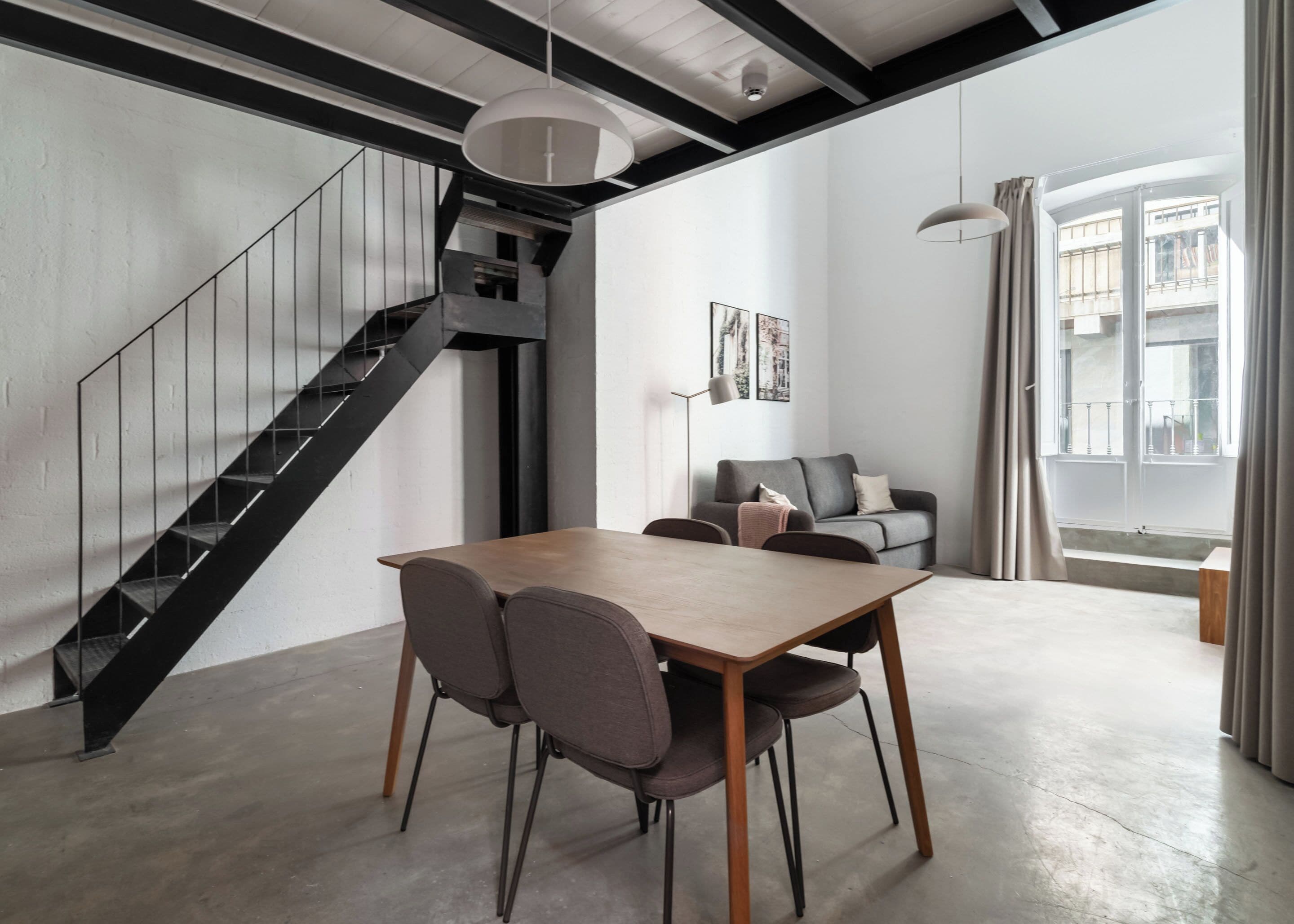 A modern interior with a wooden dining table, grey chairs, a grey sofa, a staircase, and a large window with curtains.