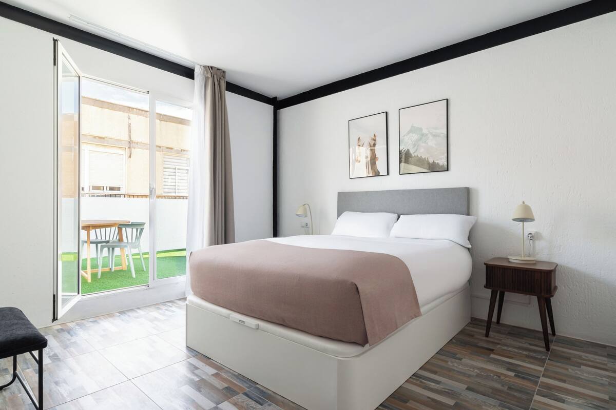 Ein Schlafzimmer mit einem Bett, einem Nachttisch, einem Sessel und Blick auf einen Außenbereich.