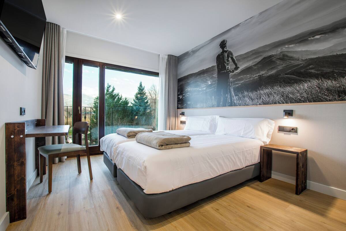 Ein modernes Schlafzimmer mit einem großen Bett, einem Schreibtisch und einem Stuhl. An der Wand hängt ein Bild und das Fenster bietet Blick auf Bäume.