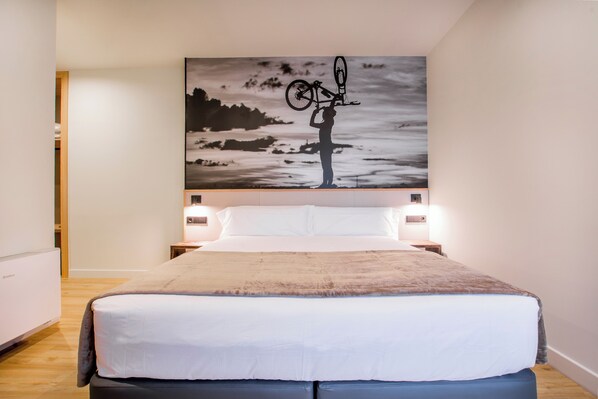 A bedroom with a large bed, a nightstand, a wall-mounted lamp, and a painting of a cyclist on a mountain.