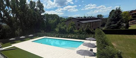 Une piscine avec une terrasse carrelée, entourée de pelouse et d’arbres, agrémentée de parasols et de chaises.
