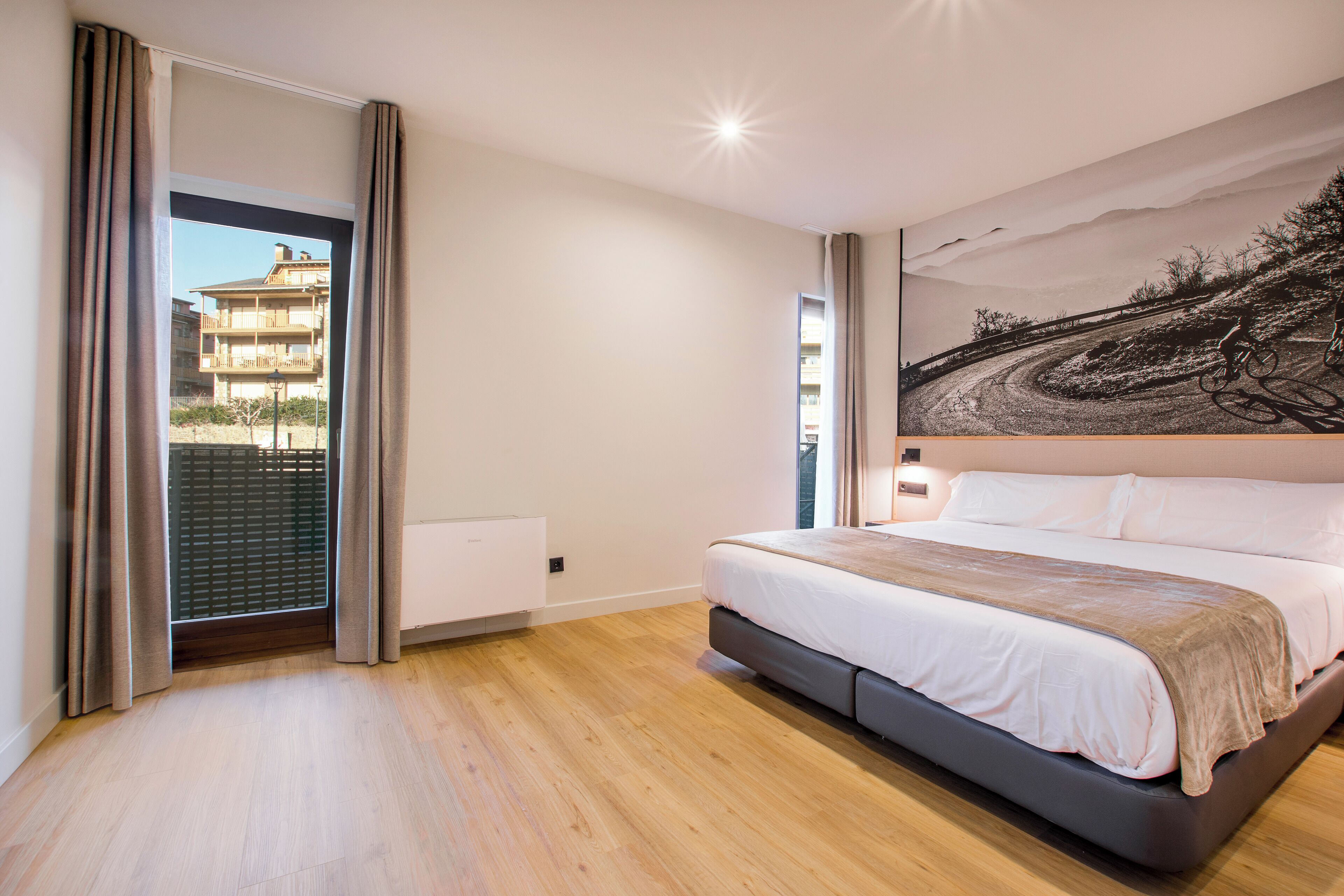 A bedroom with a large bed, a view of a building through a window, and a painting on the wall.