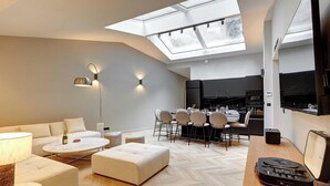 Un soggiorno moderno con divano, poggiapiedi e tavolino, una zona pranzo con sedie, una cucina con televisore e un lucernario.