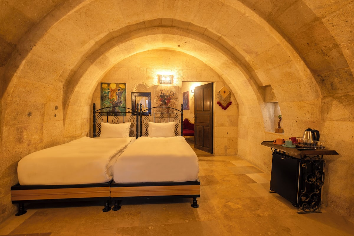 A double bed in a room with an arched stone ceiling, a small table with a kettle, and a painting on the wall.