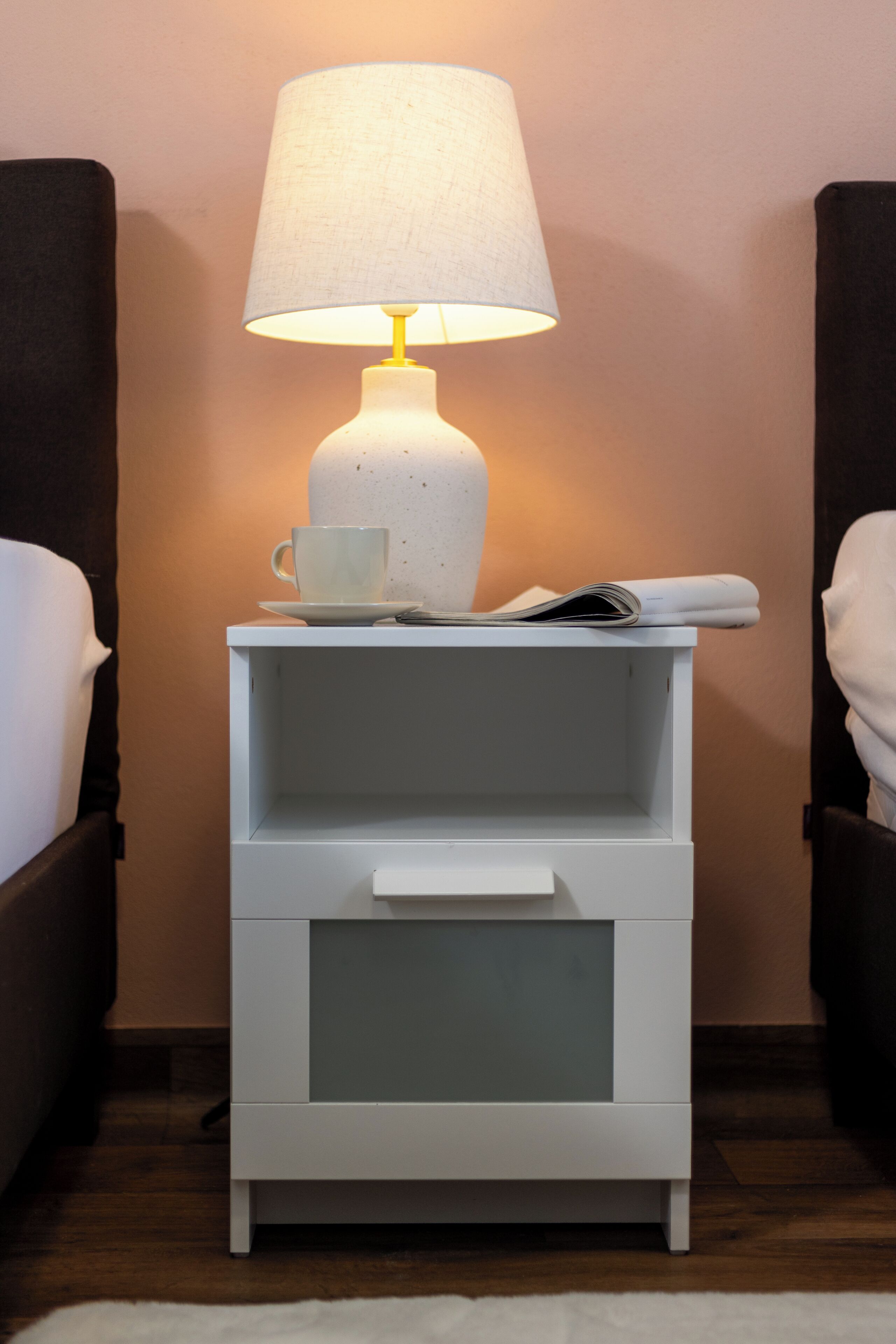 A white bedside table with a lamp, a cup, and a newspaper.